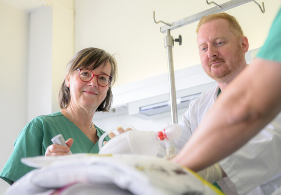 Dr. Tom Edwin Mccracken und eine HPA führen gemeinsam eine Simulation durch. Die HPA schaut dabei in die Kamera. Dr. Tom Edwin Mccracken stabilisiert ein Beatmungsgerät in der Hand.