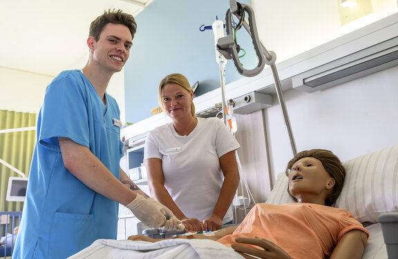 Ein HPA Auszubildener und die Hauptamtliche Praxisanleiterin stehen vor einem Krankenbett, indem eine Puppe liegt. Dabei legen die zwei Mitarbeiter ein Zugang in den Arm des Beispielpatinenten und schauen dabei in die Kamera.